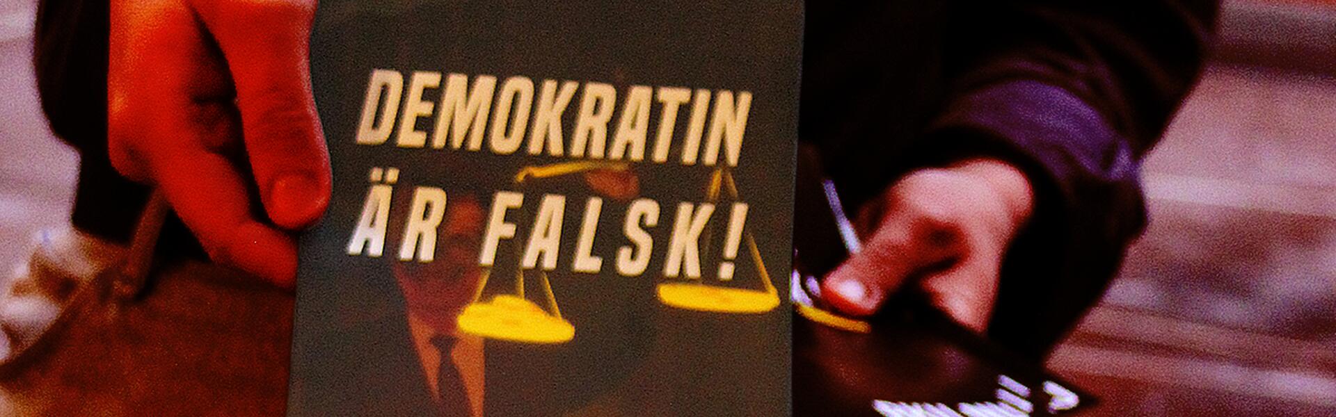 En person räcker över ett flygblad med texten "Demokratin är falsk!" på