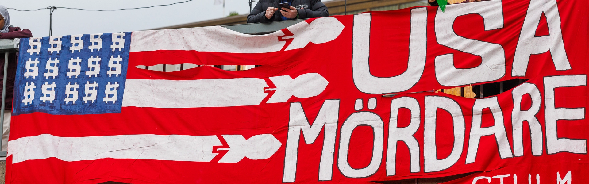 Bild p&aring; en banderoll d&auml;r det st&aring;r "USA m&ouml;rdare" och en stiliserad variant av USA:s flagga med dollartecken ist&auml;llet f&ouml;r stj&auml;rnor och komber ist&auml;llet f&ouml;r r&auml;nder.